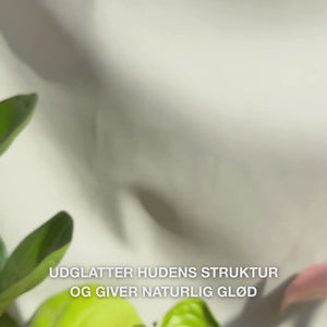 Close‑up af en hånd, der dispenserer et par tynde dråber af et gennemsigtigt, let Peeling Serum på fingerspidserne og masserer det blidt ind i ren ansigtshud — den silkelignende, flydende tekstur opløser synligt døde hudceller, forfiner hudstruktur og boostet glød med naturlige AHA (Lime Pearl), Niacinamid og Tea Tree Oil 