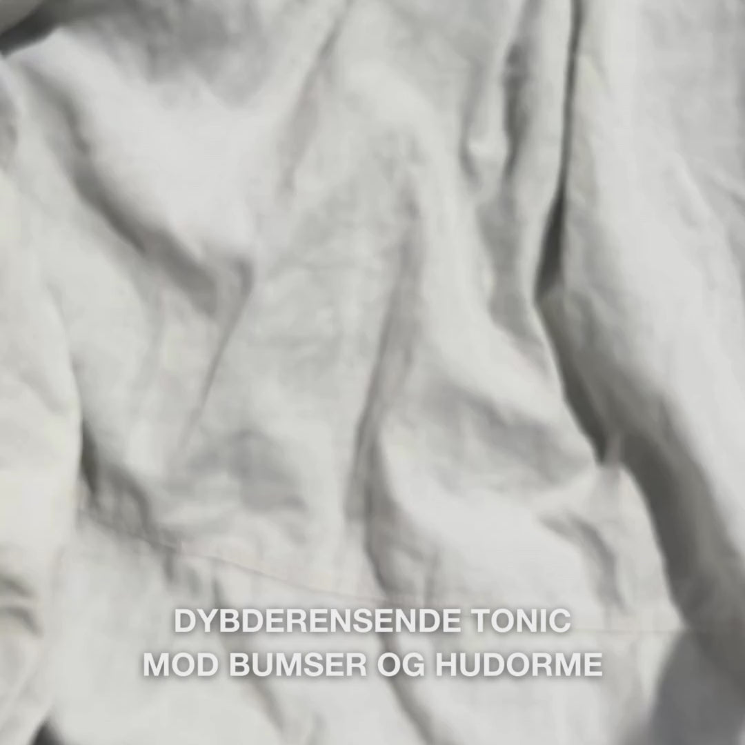 Close-up af en hånd, der forsigtigt drypper et par dråber klar, vandlignende Face Tonic med Tea Tree Oil på en vatrondel, og derefter stryger den blidt hen over ren ansigtshud: den lette, forfriskende væske beroliger, renser i dybden, fugter og efterlader huden fornyet og let forberedt til ansigtscreme.